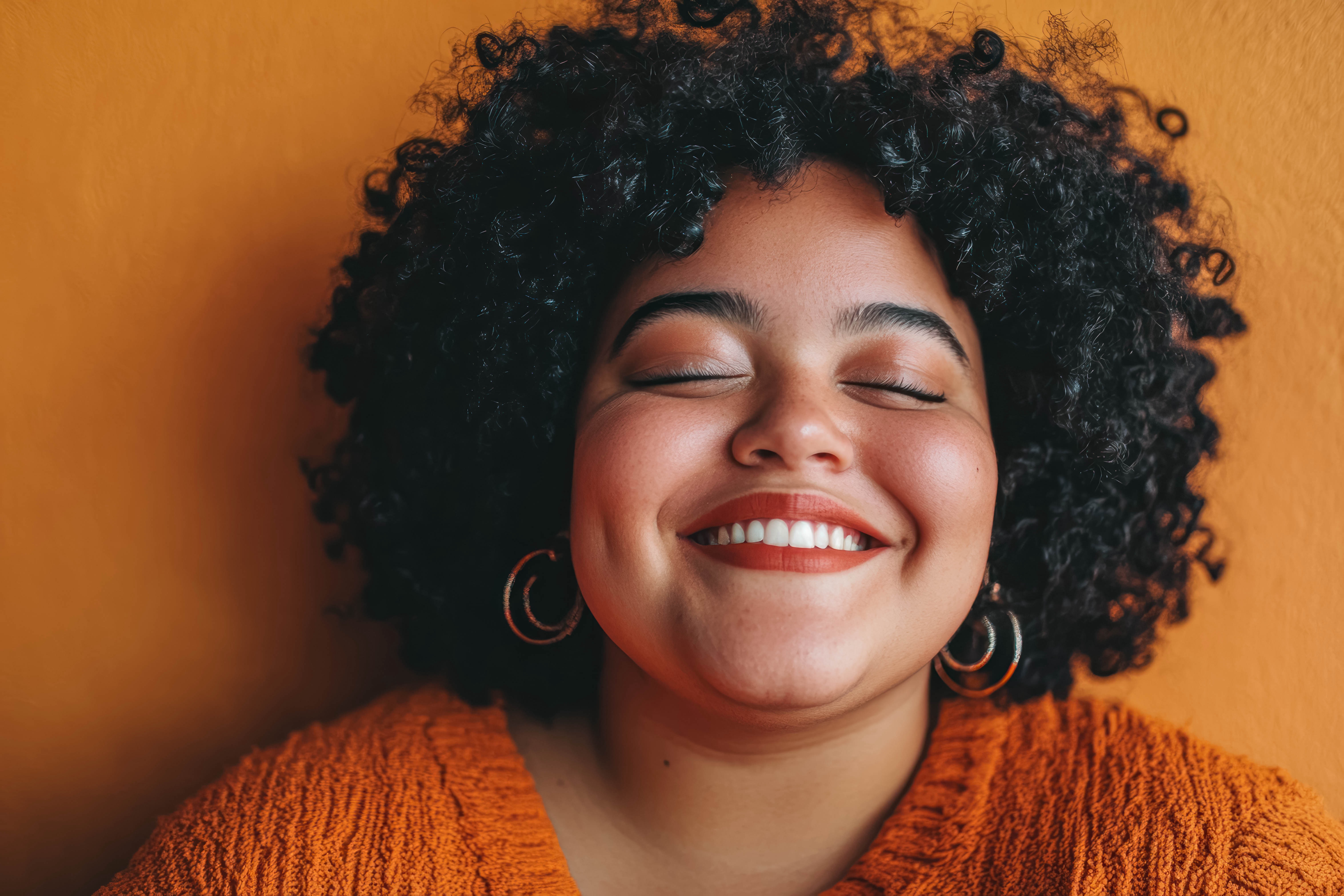 A smiling woman with natural curls and an orange sweater, eyes closed